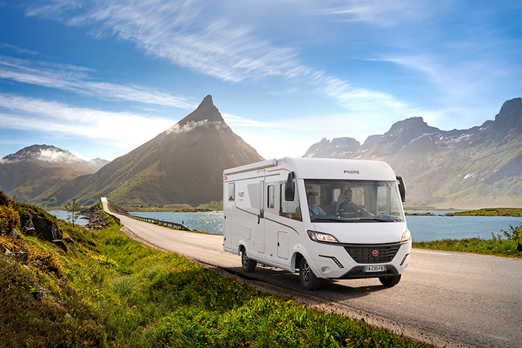 Photo d'un camping-car intégral sur une route rectiligne, à la sortie d'un lac entouré de montagnes abruptes.